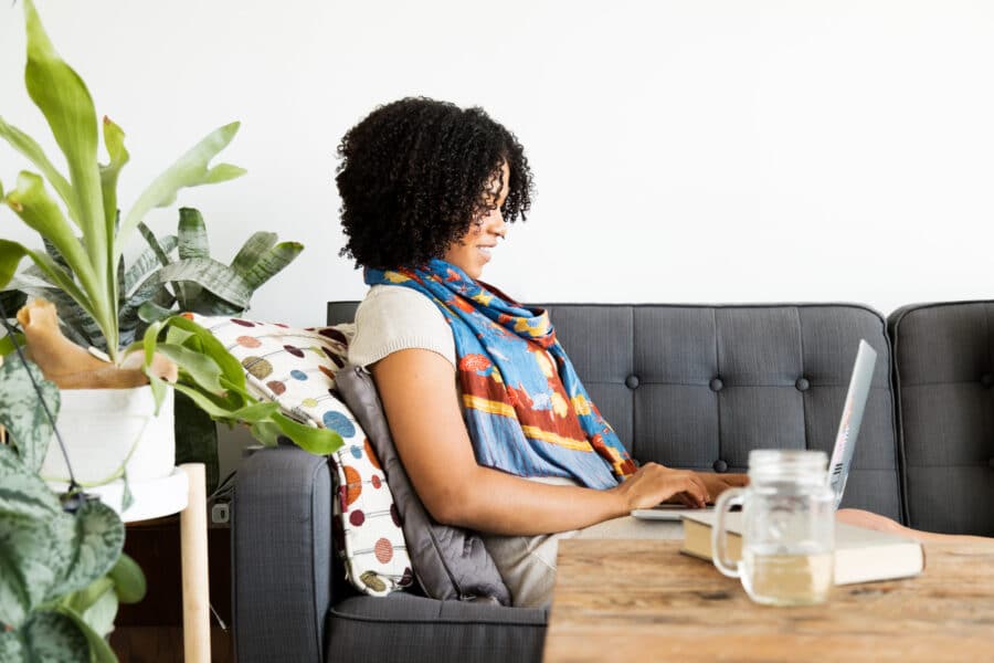 Woman sitting on a couch working on a laptop, with plants and a drink on the table.